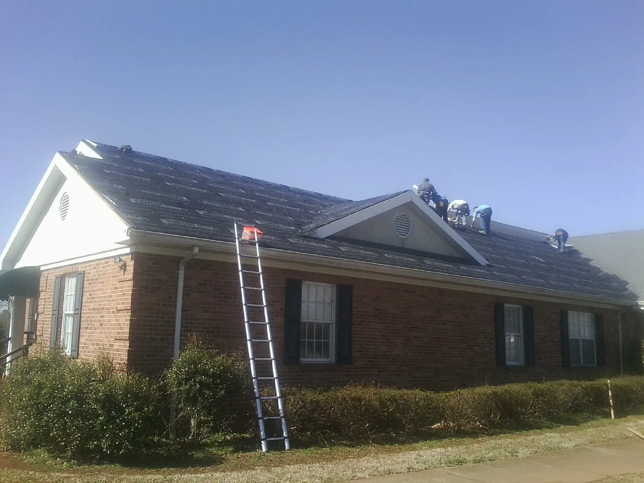 Crew tearing shingles off a brick ranch roof with a ladder set — Emergency Roof Repair work by our crew in Bronson