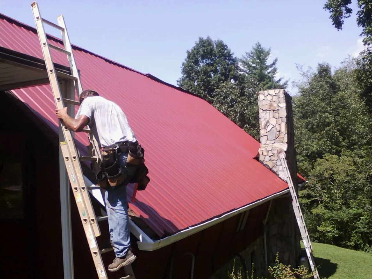 Roofer on a ladder beside a red metal roof and stone chimney — Soffit & Fascia Repair by Roofing Brothers in Bronson